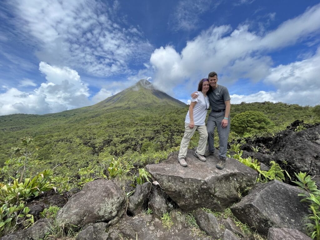 Two visitors standing on a lava boulder with the green slopes of Arenal Volcano behind