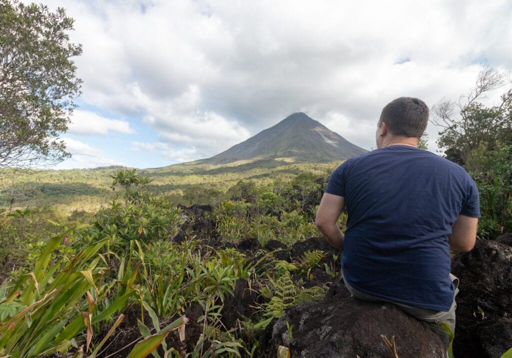 Visitor sitting on volcanic rock looking at the Arenal Volcano cone from the trail viewpoint